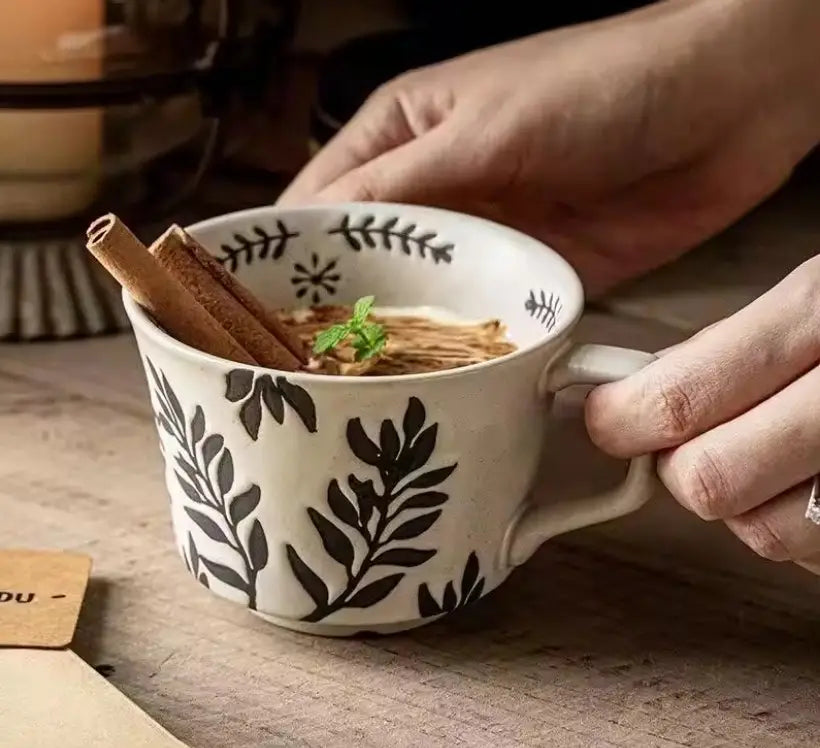 Hand holding a botanical-pattern ceramic mug with cinnamon sticks